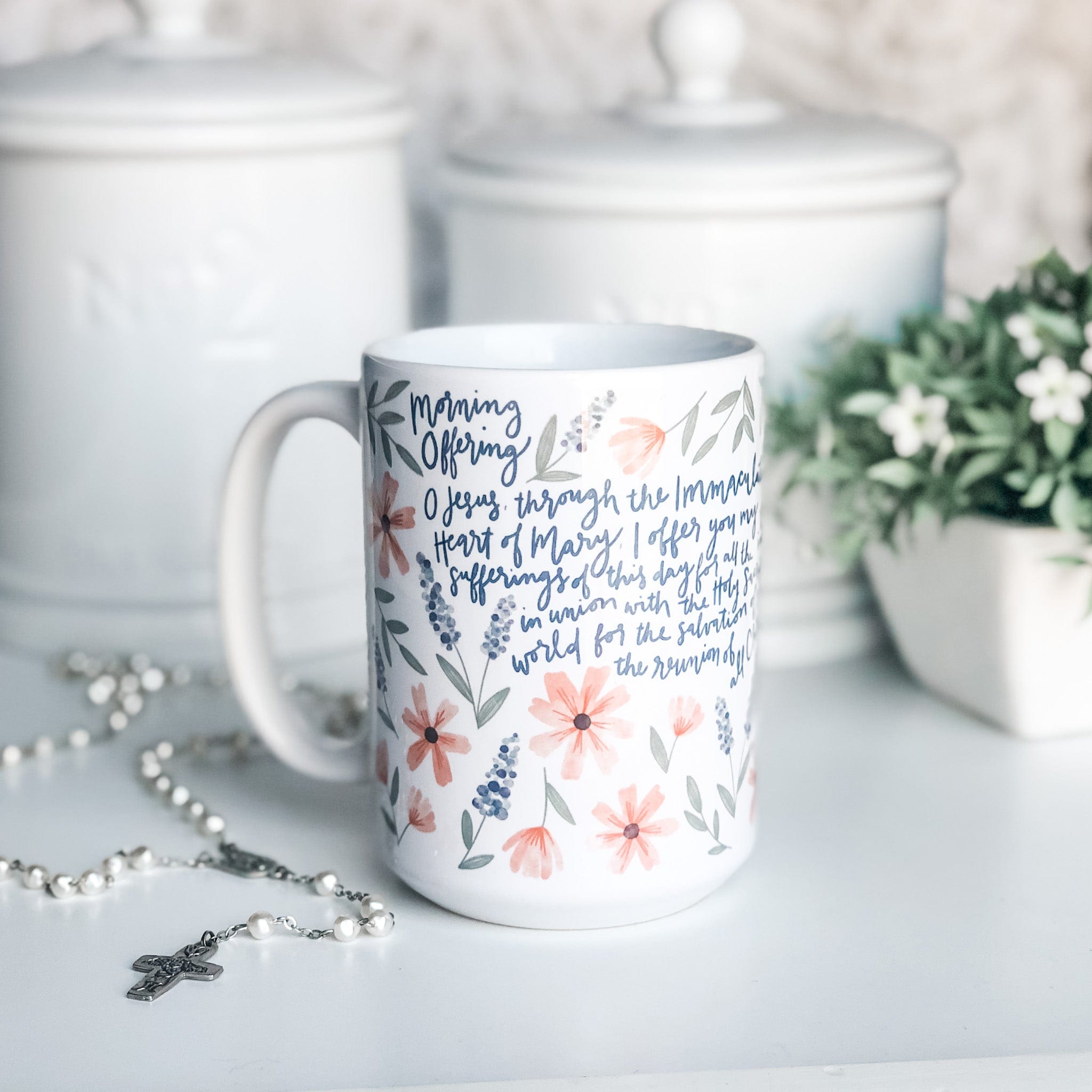 White floral mug with text, rosary, and jars.