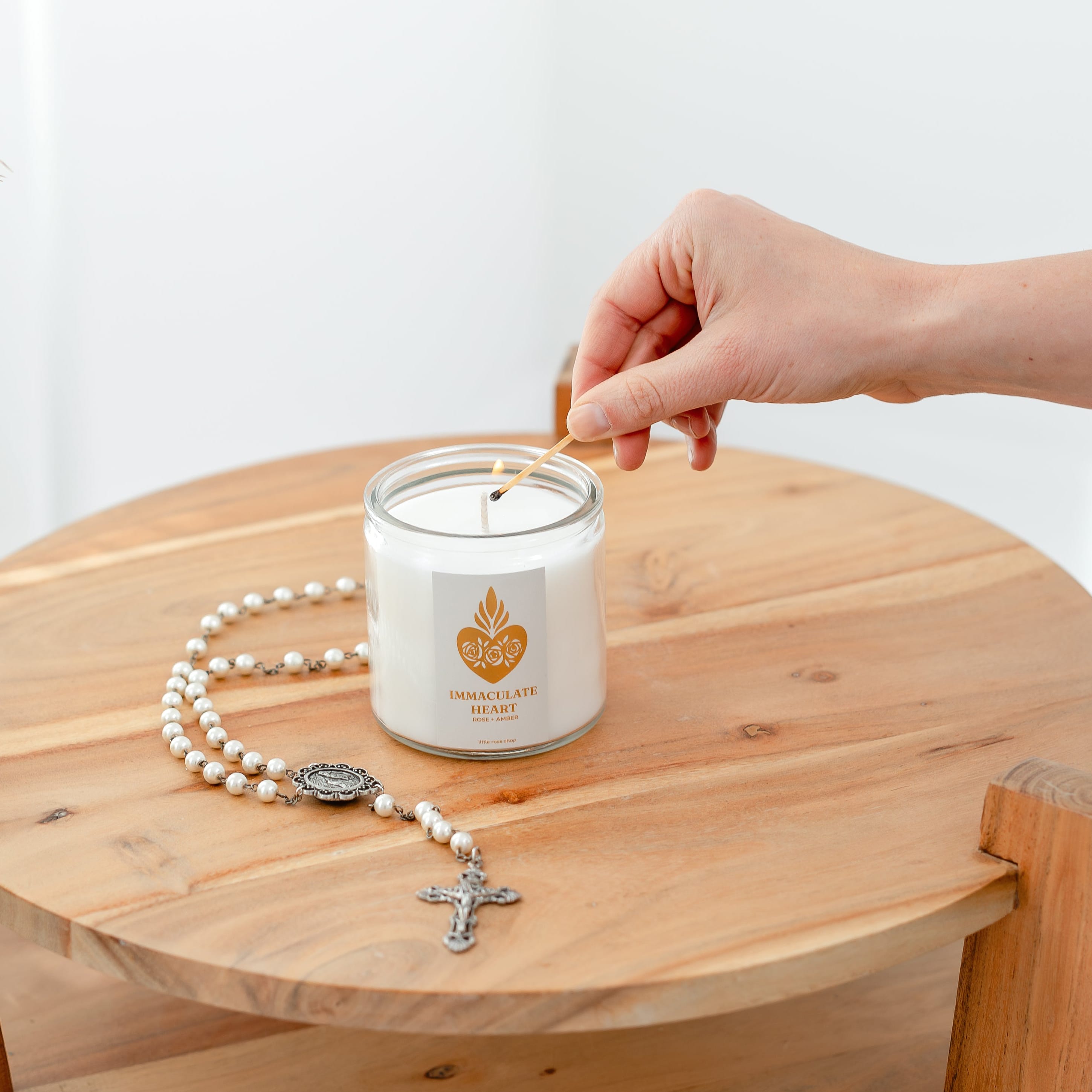 Hand placing a rosary on a candle with a wooden side table against a white background