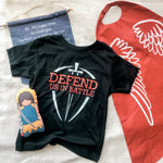 Black t-shirt with Defend Us In Battle text, cape, banner, and doll.