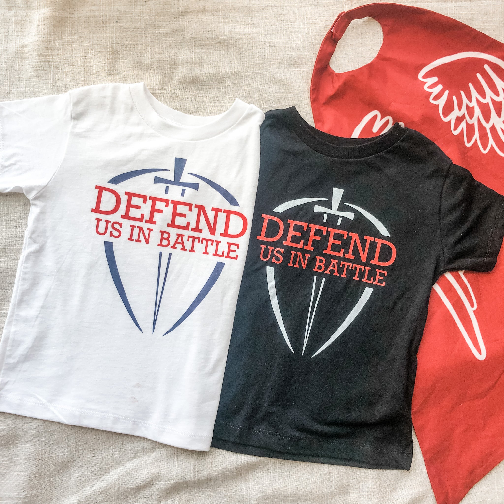 White and black shirts with Defend Us In Battle text and a red cape.